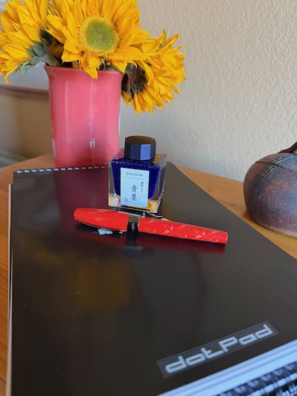 stack of Rhodia notepads with a bottle of Sailor Seiboku and Montegrappa coral colored pen in front of a vase with sunflowers